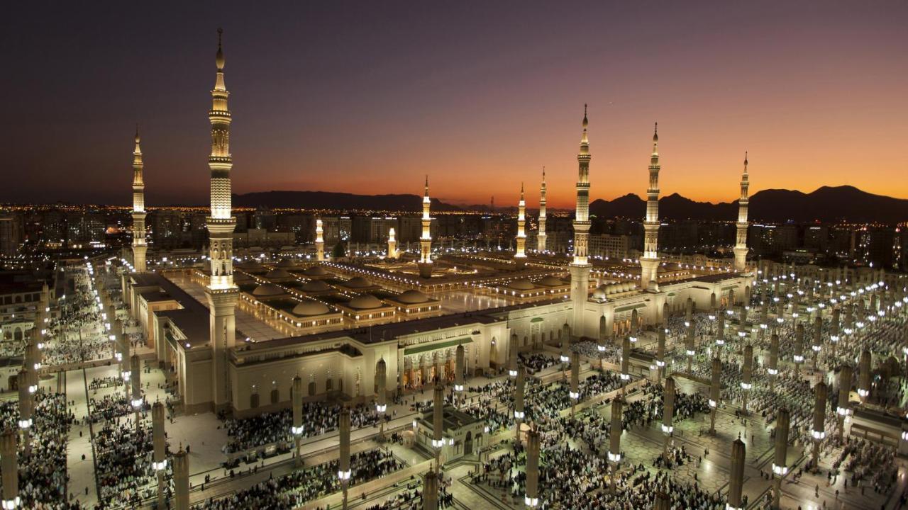 Intercontinental Dar Al Iman Madinah By Ihg Ξενοδοχείο Μεδίνα Εξωτερικό φωτογραφία The photo depicts an aerial view of a large mosque complex during sunset. The mosque features multiple tall minarets, which are intricately designed and illuminated, giving the scene a stunning appearance as the sky transitions from daylight to dusk.