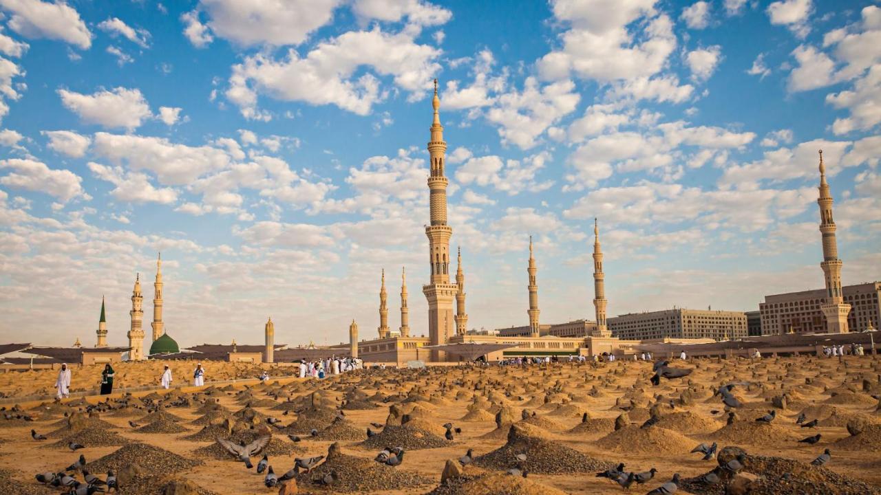Intercontinental Dar Al Iman Madinah By Ihg Ξενοδοχείο Μεδίνα Εξωτερικό φωτογραφία The photo shows a landscape that features a large mosque with a tall minaret surrounded by smaller minarets. In the foreground, there are numerous grave markers arranged in a desert-like setting. The sky is bright with scattered clouds, and there are