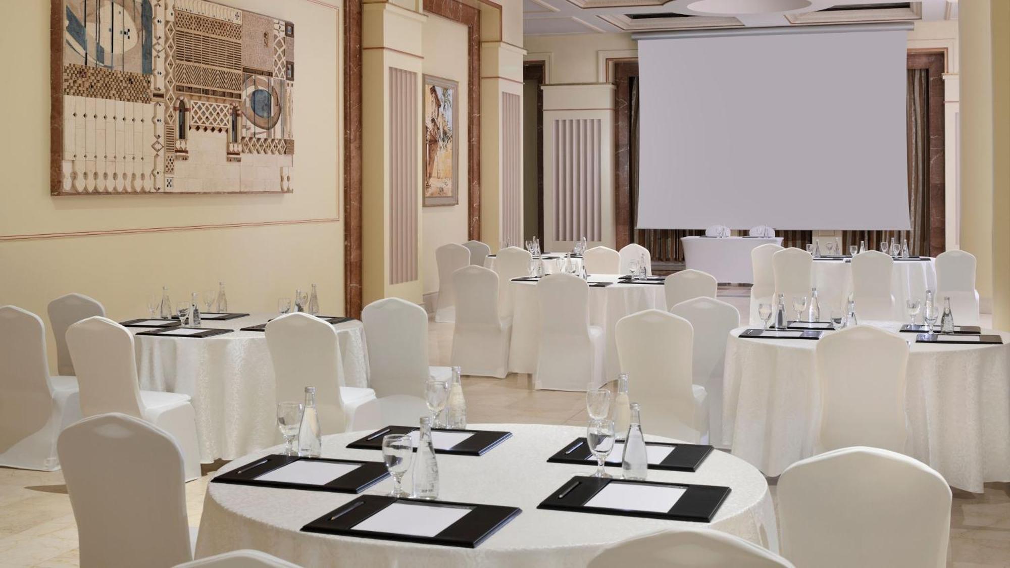 Intercontinental Dar Al Iman Madinah By Ihg Ξενοδοχείο Μεδίνα Εξωτερικό φωτογραφία The photo depicts an elegantly arranged event space. There are several round tables covered with white tablecloths and surrounded by white chairs. Each table is set with a black folder or menu and a glass of water. The walls are adorned with artworks