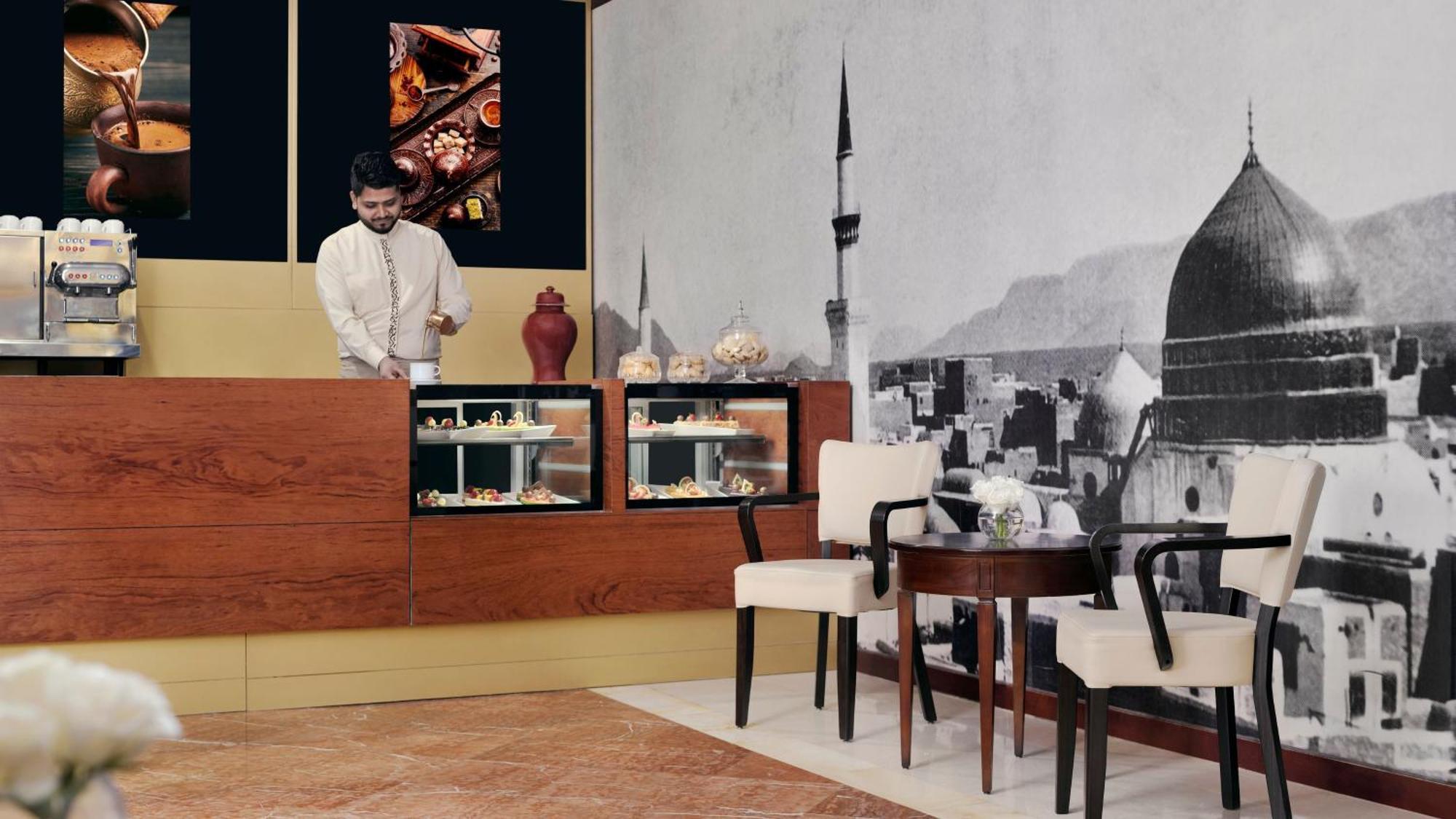 Intercontinental Dar Al Iman Madinah By Ihg Ξενοδοχείο Μεδίνα Εξωτερικό φωτογραφία The photo shows an elegant cafe or dessert shop interior. A man in traditional attire is standing behind a wooden counter, where various pastries and sweets are displayed. The walls feature a large black-and-white mural of a cityscape, possibly depic