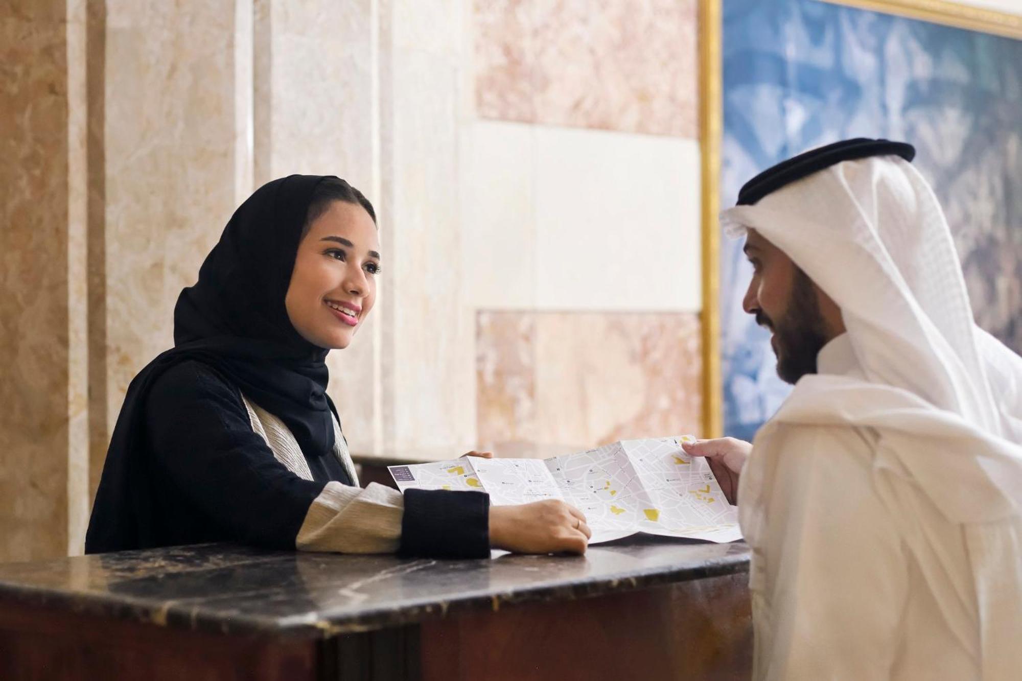 Intercontinental Dar Al Iman Madinah By Ihg Ξενοδοχείο Μεδίνα Εξωτερικό φωτογραφία The photo depicts a woman in a black hijab and a light-colored outfit smiling as she interacts with a man wearing traditional Emirati attire, including a white kandura and ghutrah (headscarf). They are at a reception desk in what appears to be a hote