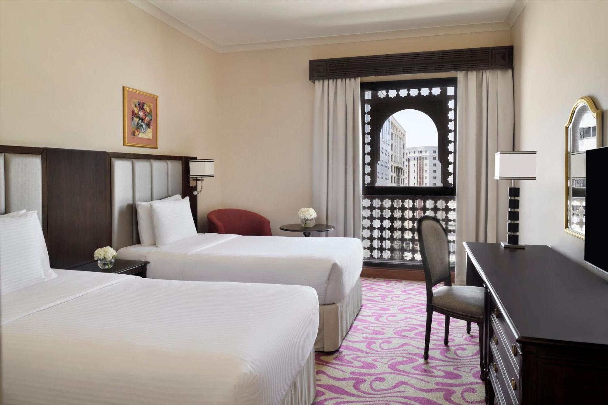 Intercontinental Dar Al Iman Madinah By Ihg Ξενοδοχείο Μεδίνα Εξωτερικό φωτογραφία The photo shows a hotel room featuring two single beds with white bedding. The room has a light, neutral color scheme and is decorated with minimal furnishings. There's a small seating area with a red chair and a round table, as well as a desk with a