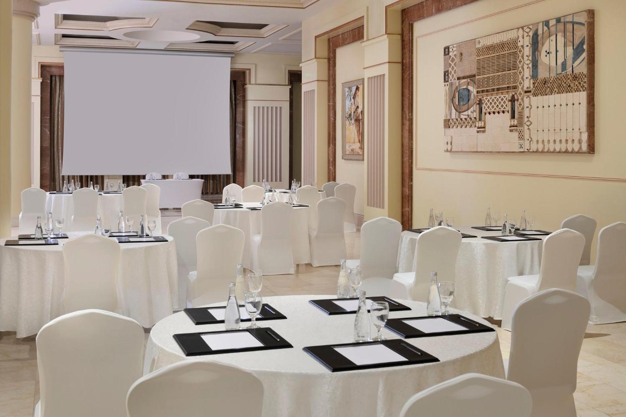 Intercontinental Dar Al Iman Madinah By Ihg Ξενοδοχείο Μεδίνα Εξωτερικό φωτογραφία The photo shows a well-decorated event space or meeting room. It features several round tables covered with white tablecloths and surrounded by white chairs. Each table has black placemats and glass water bottles, suggesting a formal setup for a gath