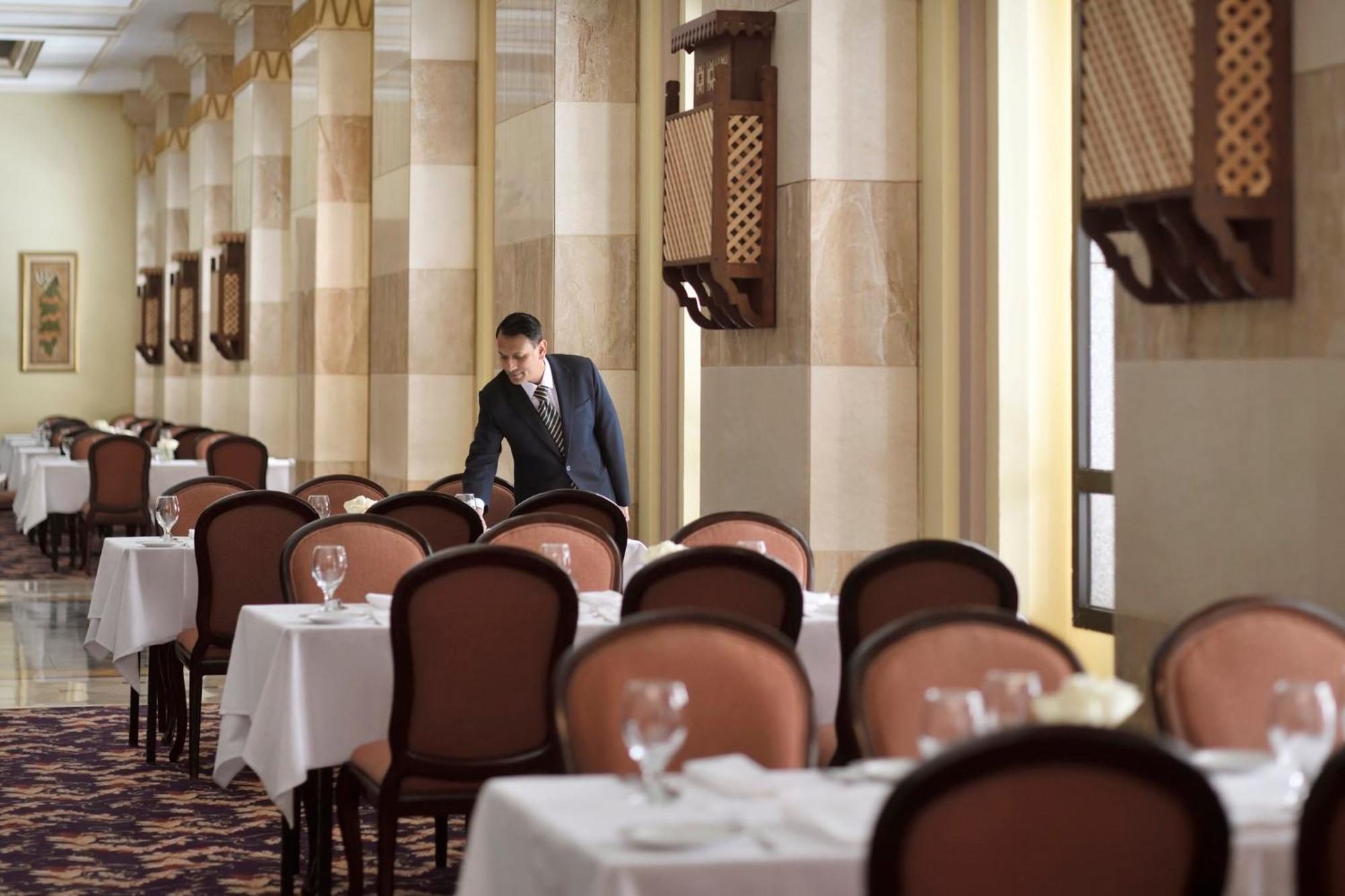 Intercontinental Dar Al Iman Madinah By Ihg Ξενοδοχείο Μεδίνα Εξωτερικό φωτογραφία The photo depicts a dining area in an elegant restaurant. There are several tables set with white tablecloths, each accompanied by chairs. A waiter, dressed in a formal suit, is seen arranging or tending to one of the tables. The restaurant features 