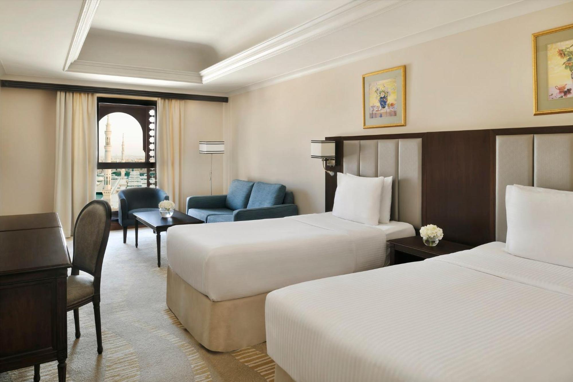 Intercontinental Dar Al Iman Madinah By Ihg Ξενοδοχείο Μεδίνα Εξωτερικό φωτογραφία The photo shows a well-appointed hotel room featuring two twin beds with white bedding. There is a small desk with a chair, a comfortable blue sofa, and a coffee table in the sitting area. The room has a warm, neutral color scheme, accented by two pa