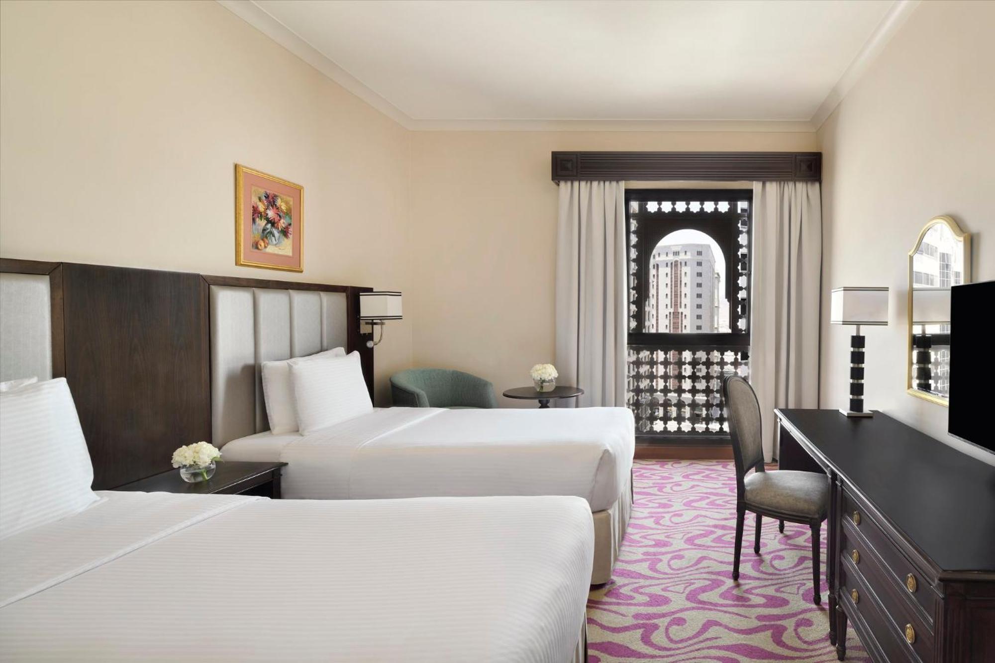 Intercontinental Dar Al Iman Madinah By Ihg Ξενοδοχείο Μεδίνα Εξωτερικό φωτογραφία The photo shows a hotel room featuring two twin beds with white bedding. There is a decorative piece of art on the wall above one of the beds. A small round table with a flower arrangement is placed next to the beds. A desk and chair are situated aga