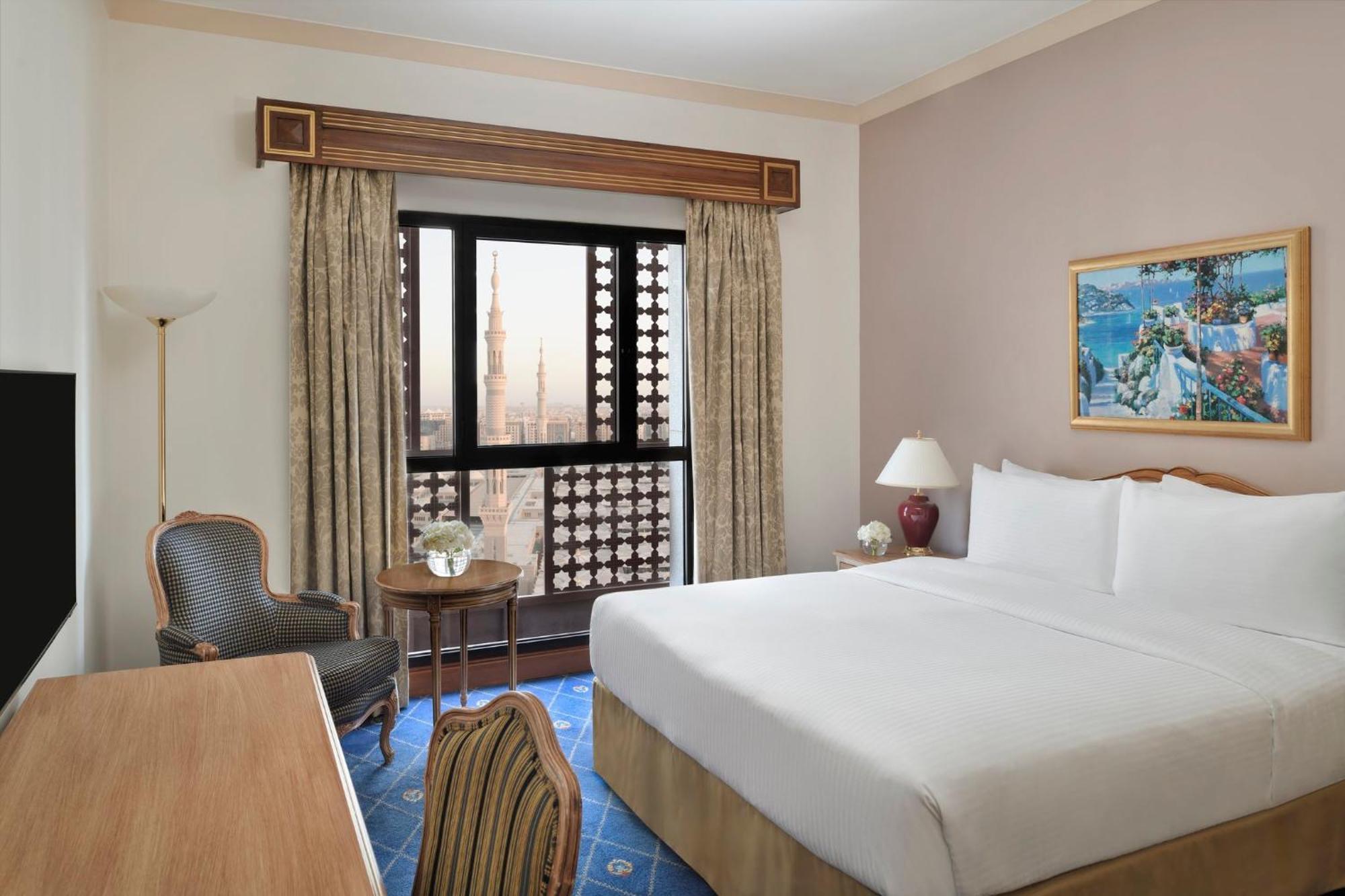 Intercontinental Dar Al Iman Madinah By Ihg Ξενοδοχείο Μεδίνα Εξωτερικό φωτογραφία The photo shows a hotel room with a modern design. There is a large bed with white linens placed against the wall. Next to the bed, there is a small bedside table with a lamp. A comfortable armchair is situated nearby, accompanied by a wooden desk. T