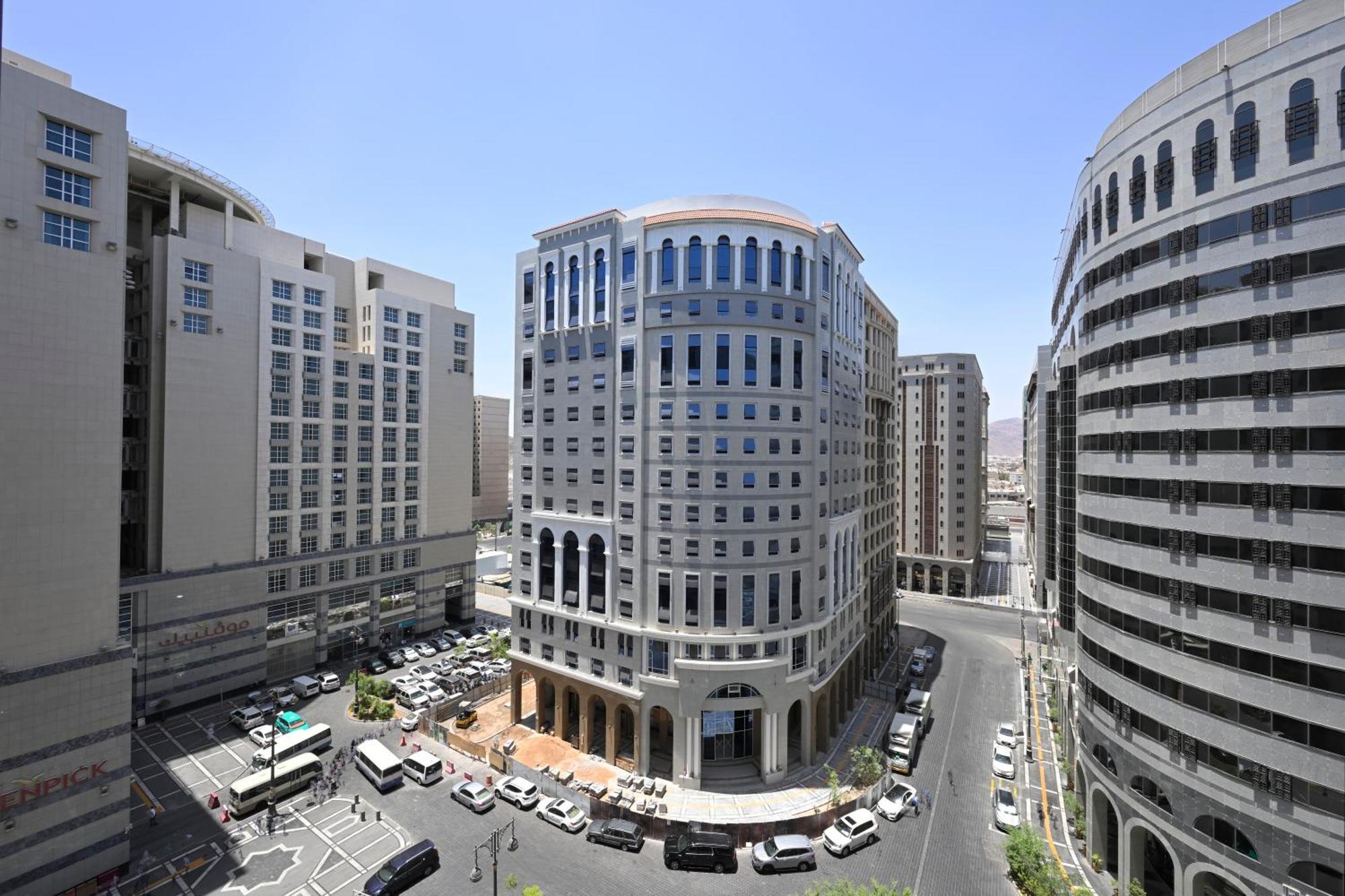 Intercontinental Dar Al Iman Madinah By Ihg Ξενοδοχείο Μεδίνα Εξωτερικό φωτογραφία The photo shows a modern urban scene featuring several buildings, including a prominent rounded building with a glass façade. This building appears to be situated at an intersection, surrounded by other high-rise structures that have distinct archite