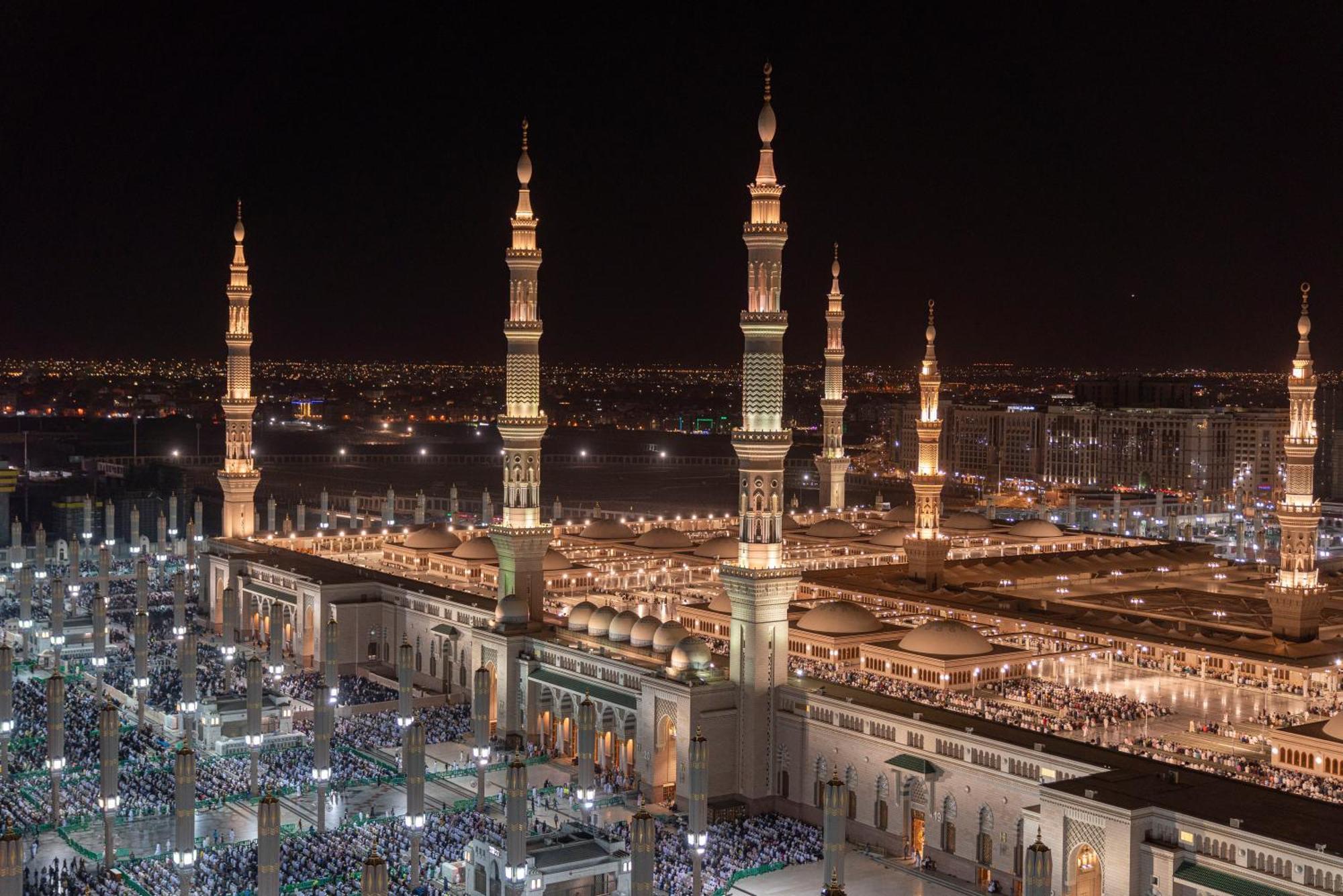 Intercontinental Dar Al Iman Madinah By Ihg Ξενοδοχείο Μεδίνα Εξωτερικό φωτογραφία The photo depicts a stunning nighttime view of a large mosque complex, prominently featuring tall minarets that are illuminated against the dark sky. The surrounding area is bustling with people, likely worshippers, gathered in the courtyard of the m