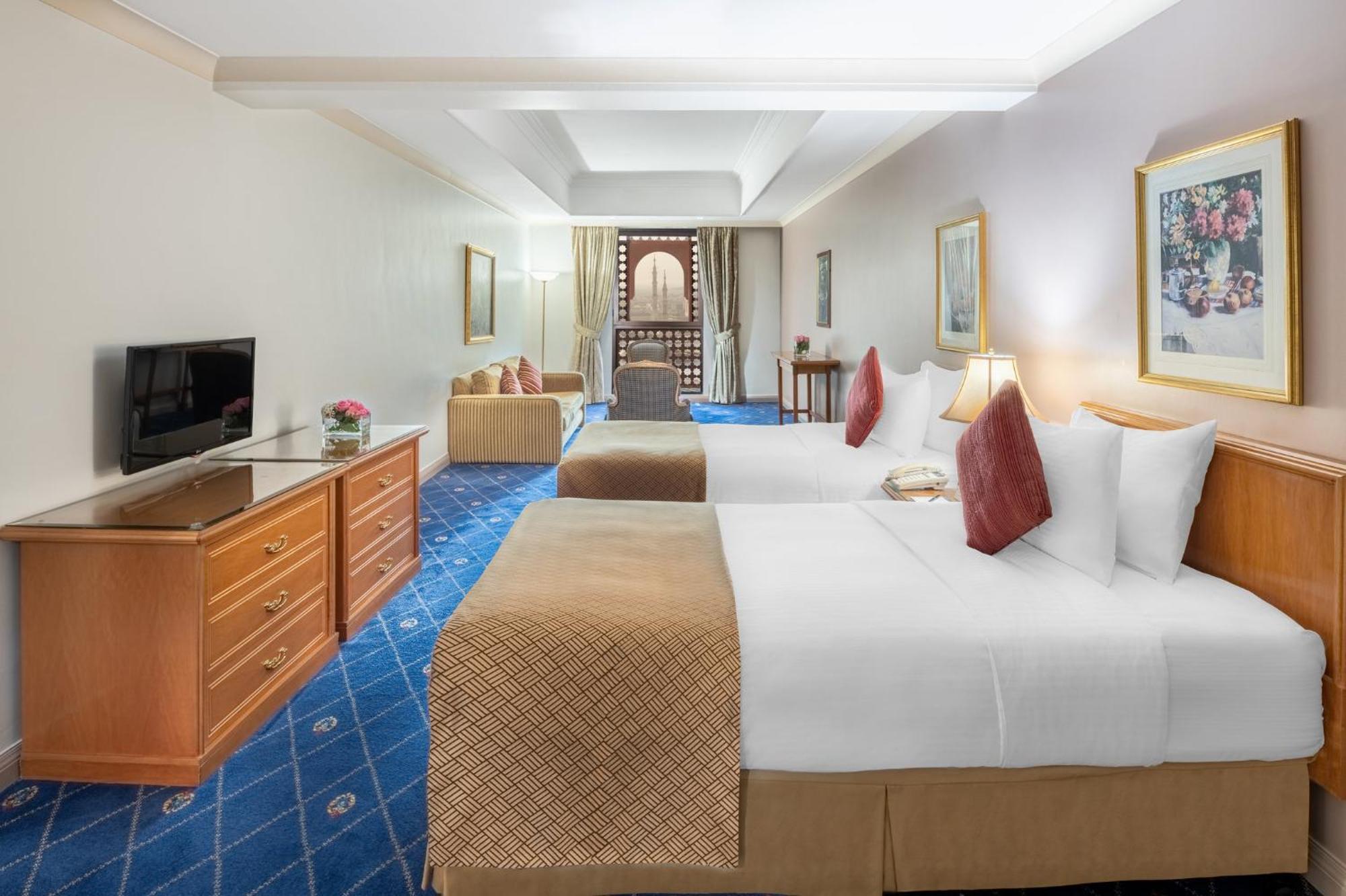 Intercontinental Dar Al Iman Madinah By Ihg Ξενοδοχείο Μεδίνα Εξωτερικό φωτογραφία The photo shows a hotel room featuring two beds with white linens and decorative pillows. There is a wooden dresser with a television on it. The flooring has a blue carpet with a pattern, and the walls are painted in a light color. The room is well-l