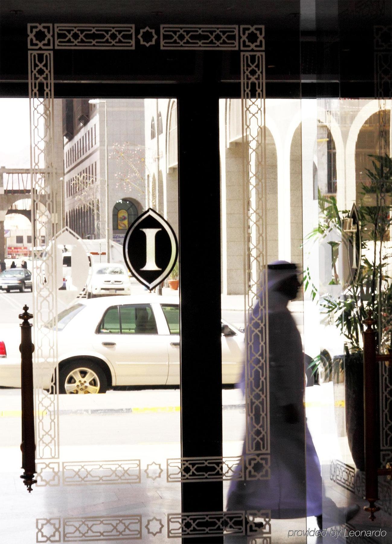 Intercontinental Dar Al Iman Madinah By Ihg Ξενοδοχείο Μεδίνα Εσωτερικό φωτογραφία The photo shows a view through a glass door or window, featuring an elegant design with a prominent "I" symbol displayed. Through the glass, there are some parked vehicles visible, including a white car. In the foreground, a shadowy figure of a perso
