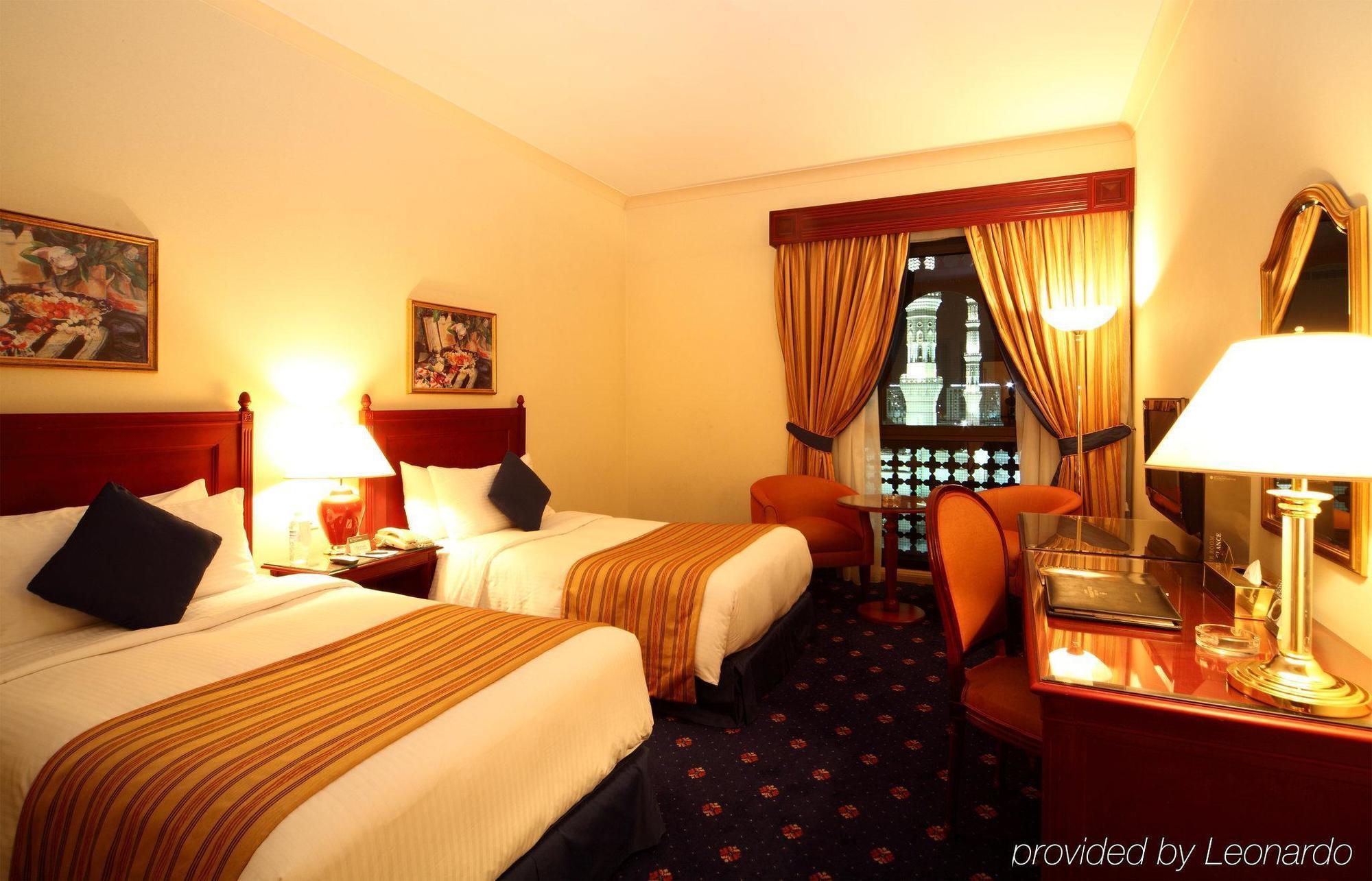 Intercontinental Dar Al Iman Madinah By Ihg Ξενοδοχείο Μεδίνα Δωμάτιο φωτογραφία The photo shows a hotel room featuring two twin beds with white linens and brown striped bedspreads. There are decorative pillows on the beds, and the room has warm-colored walls. To the side, there's a small sitting area with a round table and a cha