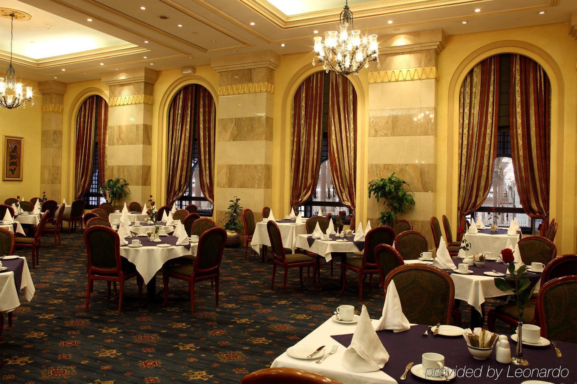Intercontinental Dar Al Iman Madinah By Ihg Ξενοδοχείο Μεδίνα Εστιατόριο φωτογραφία The photo depicts an elegant restaurant interior. The space features ornate decor with tall windows draped in rich, patterned curtains. Tables are neatly set with white tablecloths and napkins, and there are plush chairs arranged around them. The flo