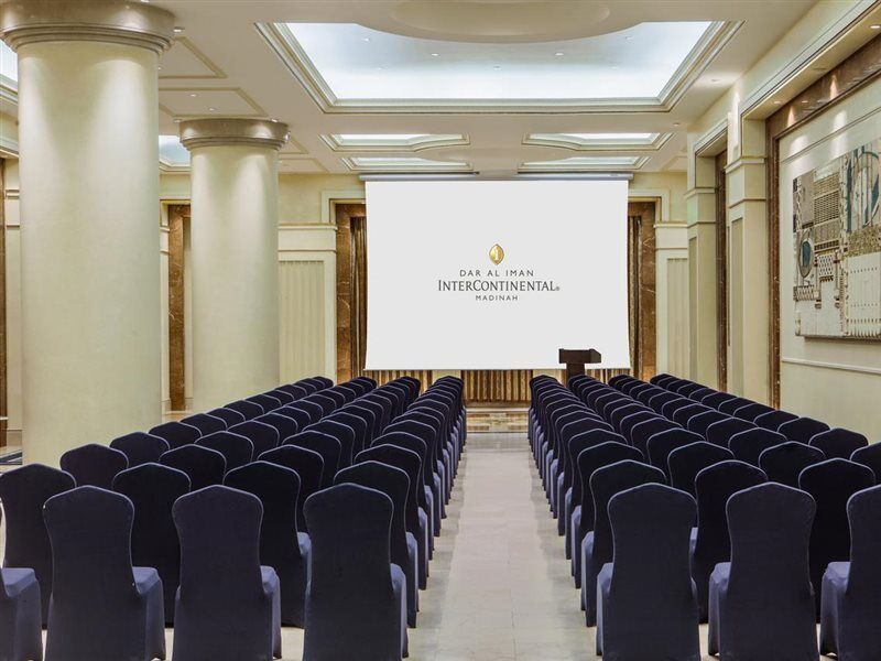 Intercontinental Dar Al Iman Madinah By Ihg Ξενοδοχείο Μεδίνα Εξωτερικό φωτογραφία The photo shows a conference or meeting room set up. There are rows of dark blue chairs arranged facing a large screen at the front of the room. The screen displays the logo of the Intercontinental hotel, indicating that this space is likely intended