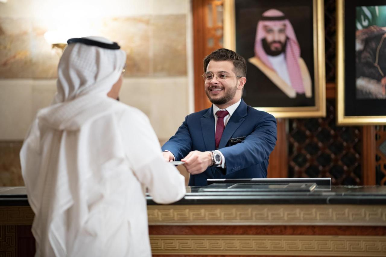 Intercontinental Dar Al Iman Madinah By Ihg Ξενοδοχείο Μεδίνα Εξωτερικό φωτογραφία The photo shows a hotel reception area. A male receptionist, dressed in a navy suit with a tie, is smiling and handing an item to a guest who is wearing traditional white attire, possibly a kandura or thobe. The background features elegantly decorate