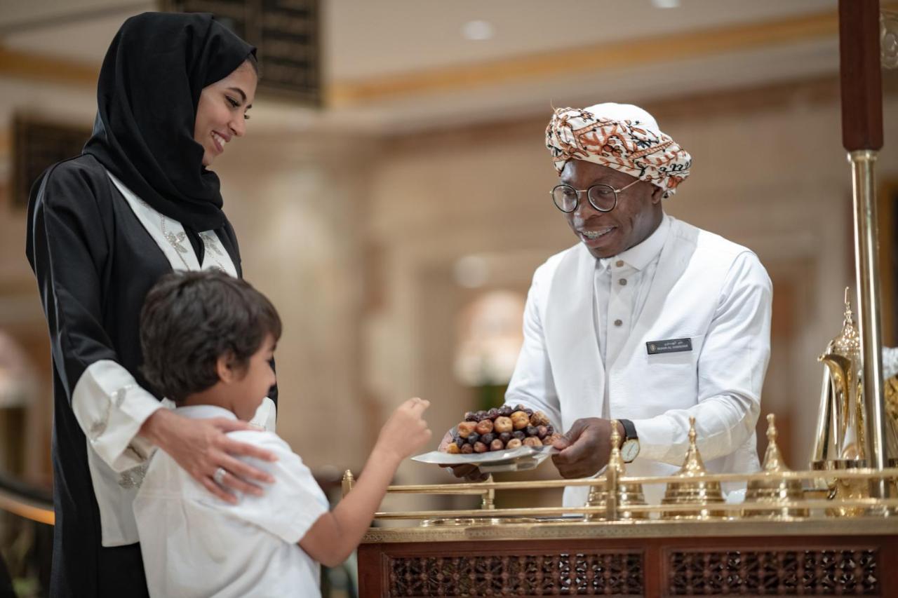 Intercontinental Dar Al Iman Madinah By Ihg Ξενοδοχείο Μεδίνα Εξωτερικό φωτογραφία The photo depicts a warm interaction between three people. A woman, dressed in a traditional black attire and a headscarf, stands beside a young boy who is reaching out towards a plate of dates held by a smiling man in a white traditional outfit with